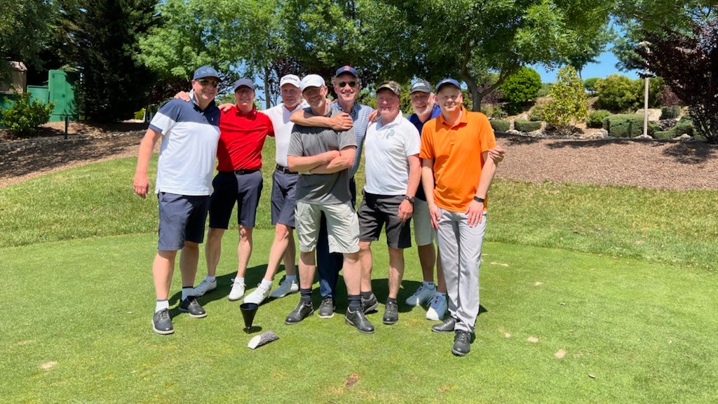 Otte mænd står sammen og poserer smilende på en golfbane, omgivet af grønt græs og træer i baggrunden.