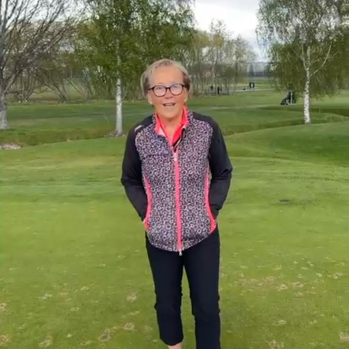 Ældre kvinde med briller står smilende på en golfbane med grønt græs og træer i baggrunden.
