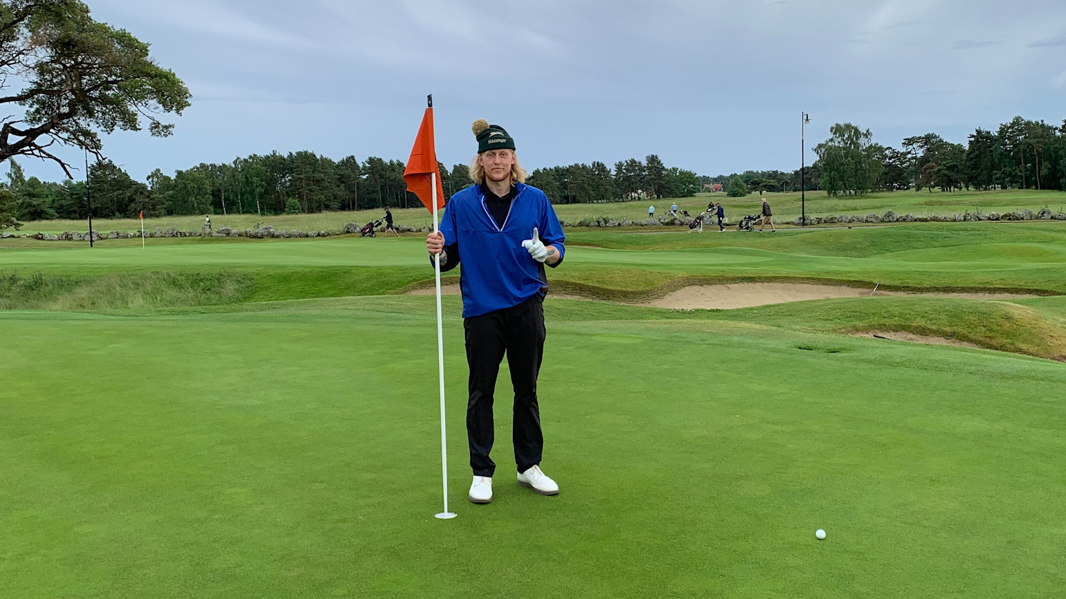 Henkilö, jolla on sininen paita ja pipo, pitää kiinni lipputangosta golfviheriöllä, näyttää yhtä sormea ja pitelee golfpalloa, taustalla näkyy golfkenttä.