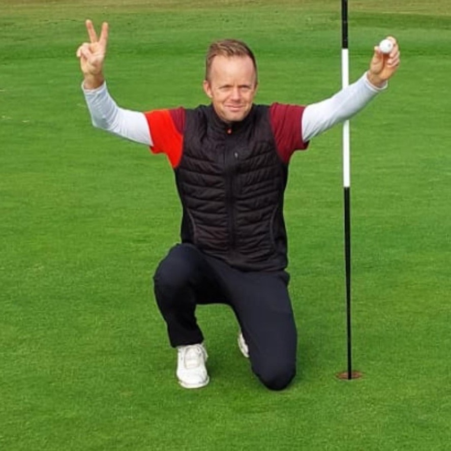 Man knæler på en golfbane, holder en golfbold op og laver sejrtegn ved siden af et golfhul med flag.