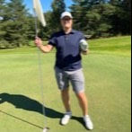 En person står på en golfbane, holder en flagstang i den ene hånd og en golfbold i den anden, omgivet af grønne omgivelser.