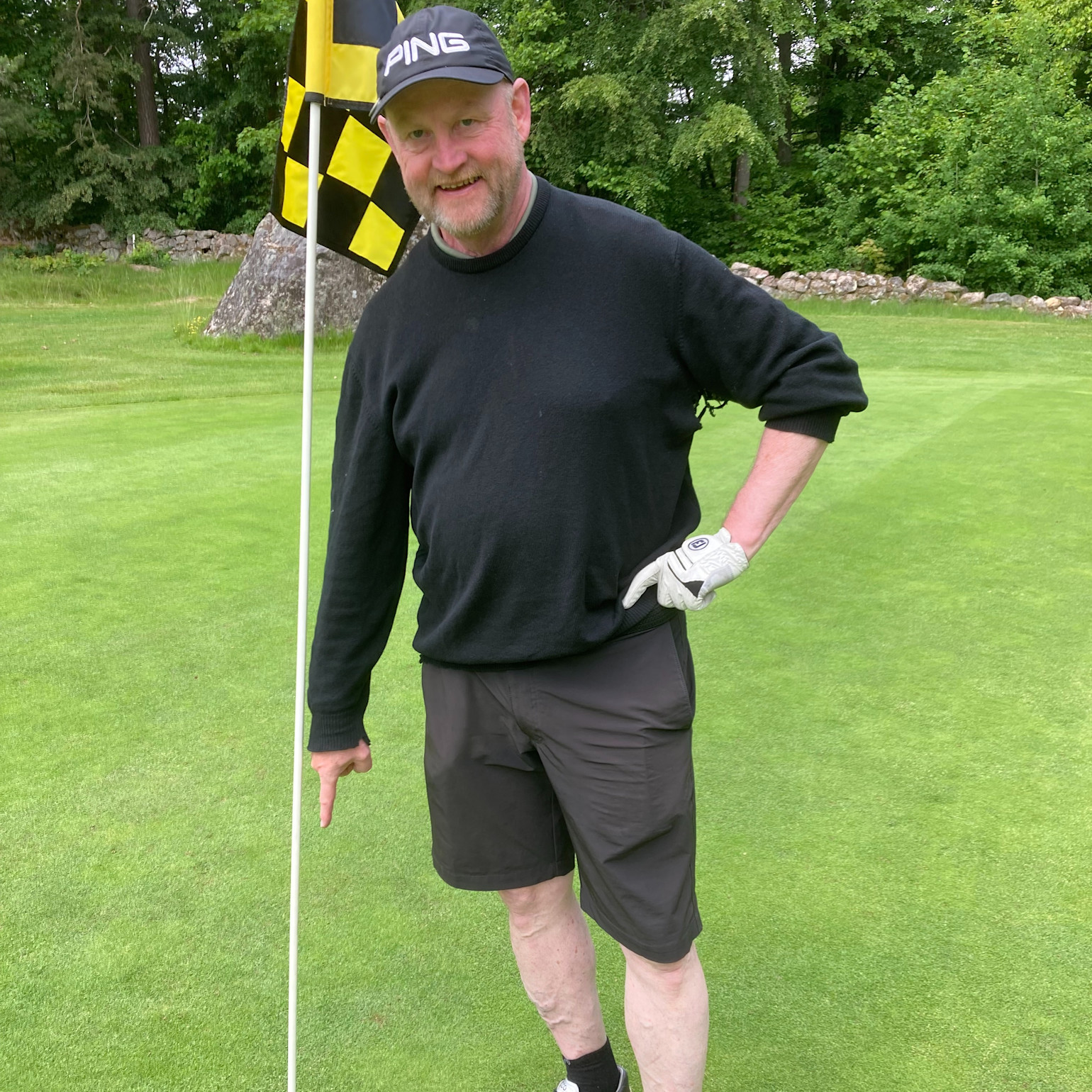 Mand iført sort trøje og kasket med teksten "PING" peger mod jorden ved siden af et golf-flag på en grøn golfbane.