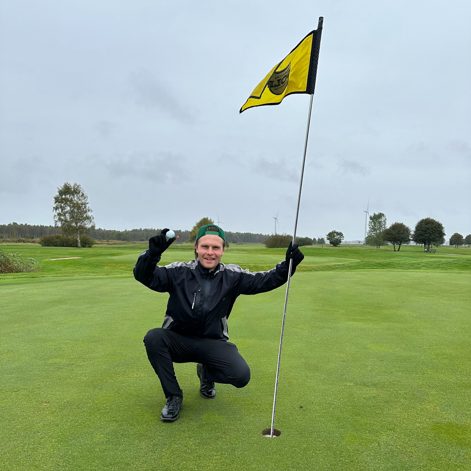 Mies urheiluvaatteissa kyykistyy golfreiän vieressä, pitää golfpalloa toisessa kädessä ja lippukeppiä toisessa kädessä golfkentällä.