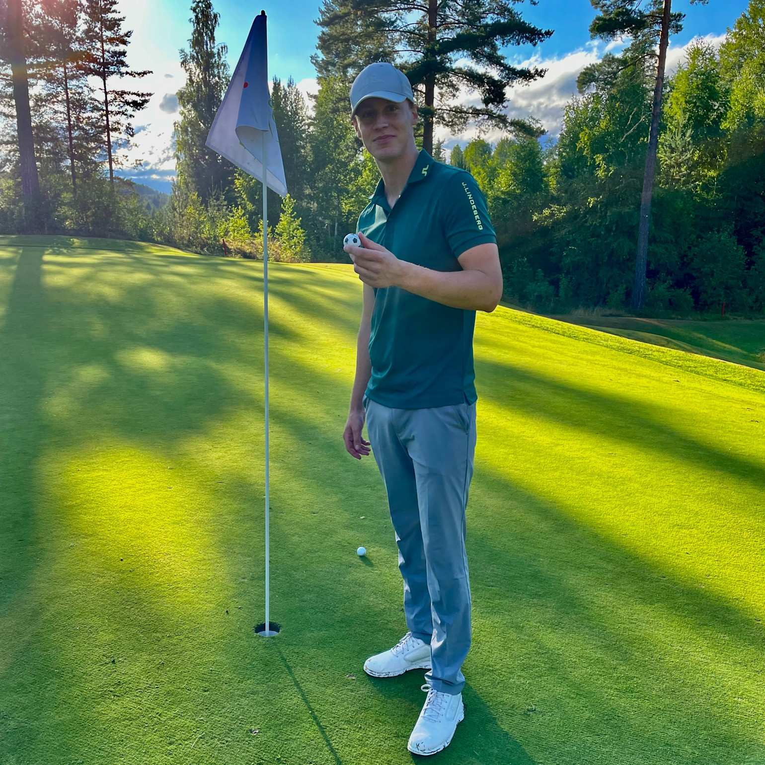 En mand i golfbeklædning holder en golfbold ved siden af et hul på en solrig golfbane omgivet af træer.