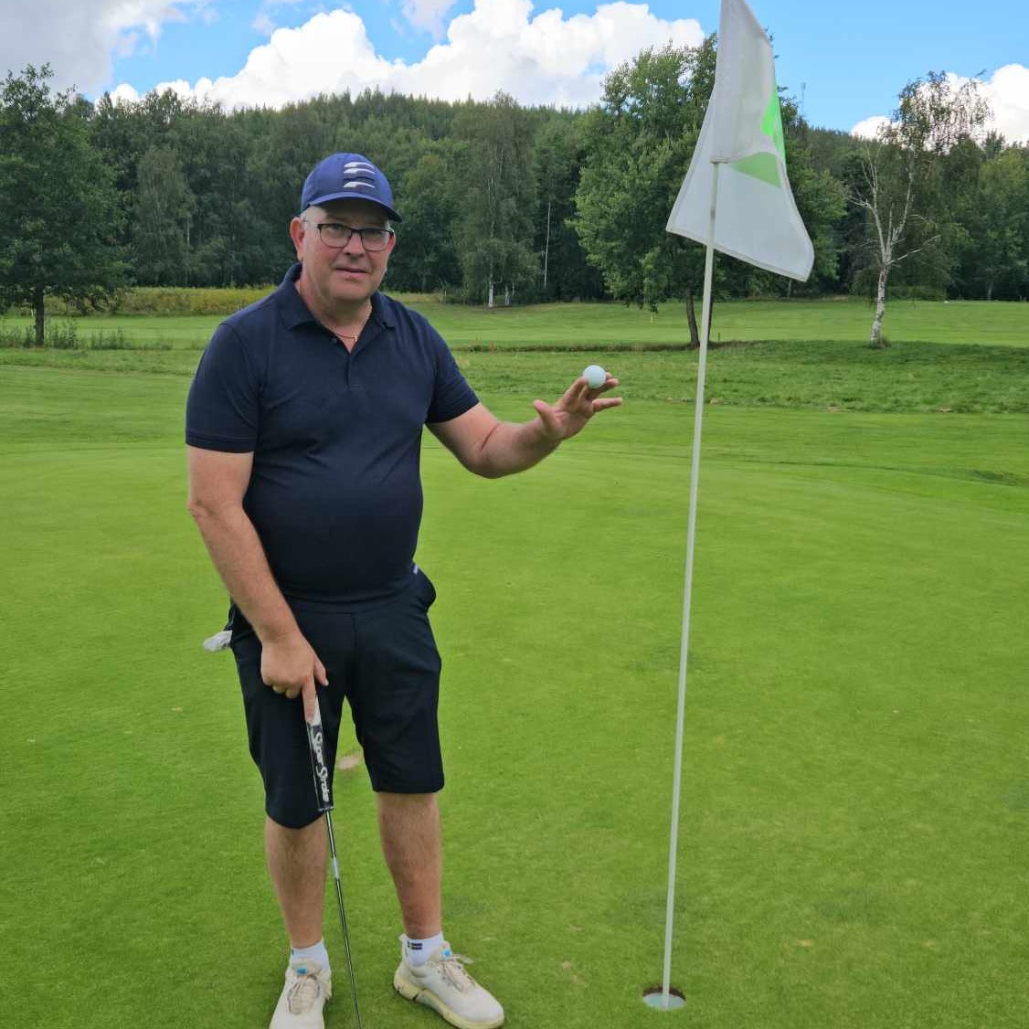 Man i mörkblå golfkläder håller en golfboll bredvid hålet på en golfbana, med skog i bakgrunden.