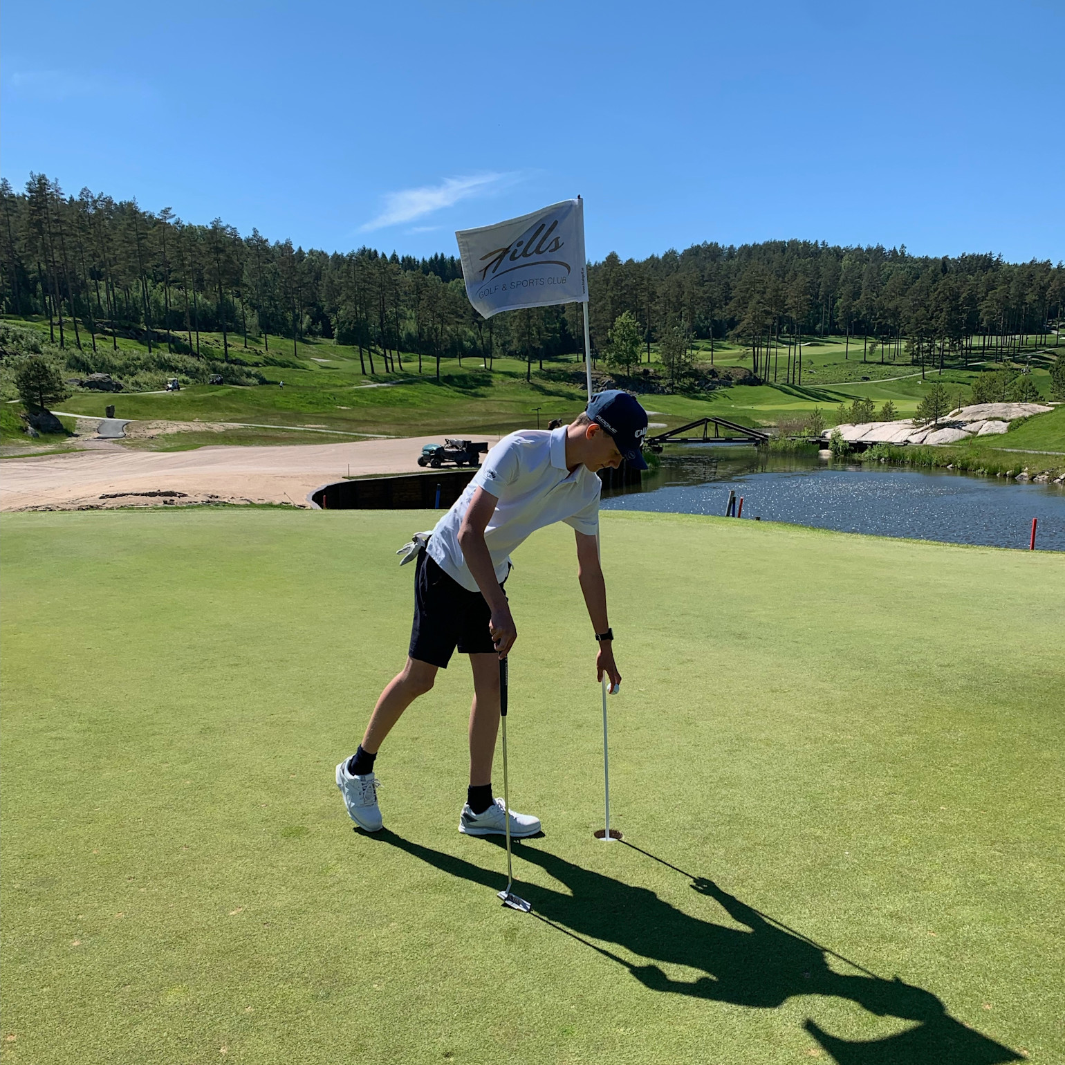 En golfspiller i hvid trøje og mørke shorts samler en bold op fra hullet på en grøn bane med flag, sø og skov i baggrunden.