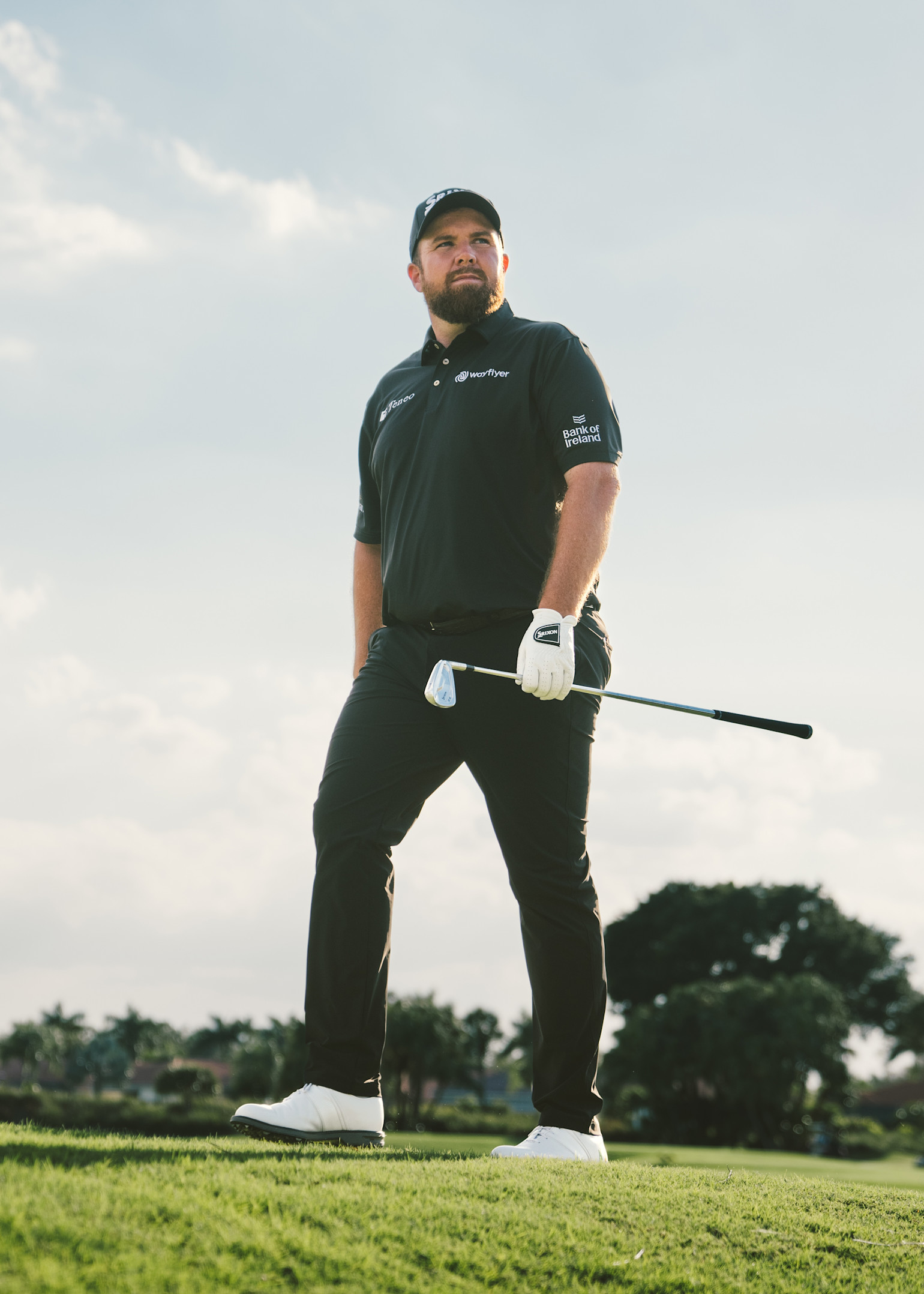 Ein Mann in schwarzer Golfkleidung und mit einer Kappe steht auf einem Golfplatz mit einem Golfschläger in der Hand, Bäume im Hintergrund.