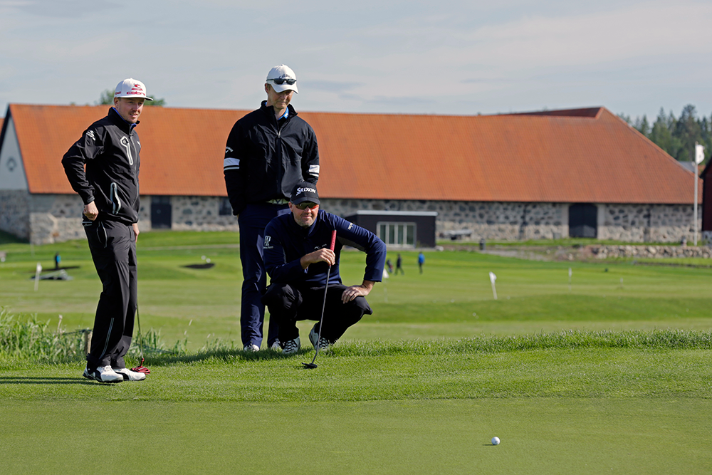 Tre golfspelare står och hukar vid en green och tittar på en golfboll, med en stenlada i bakgrunden.
