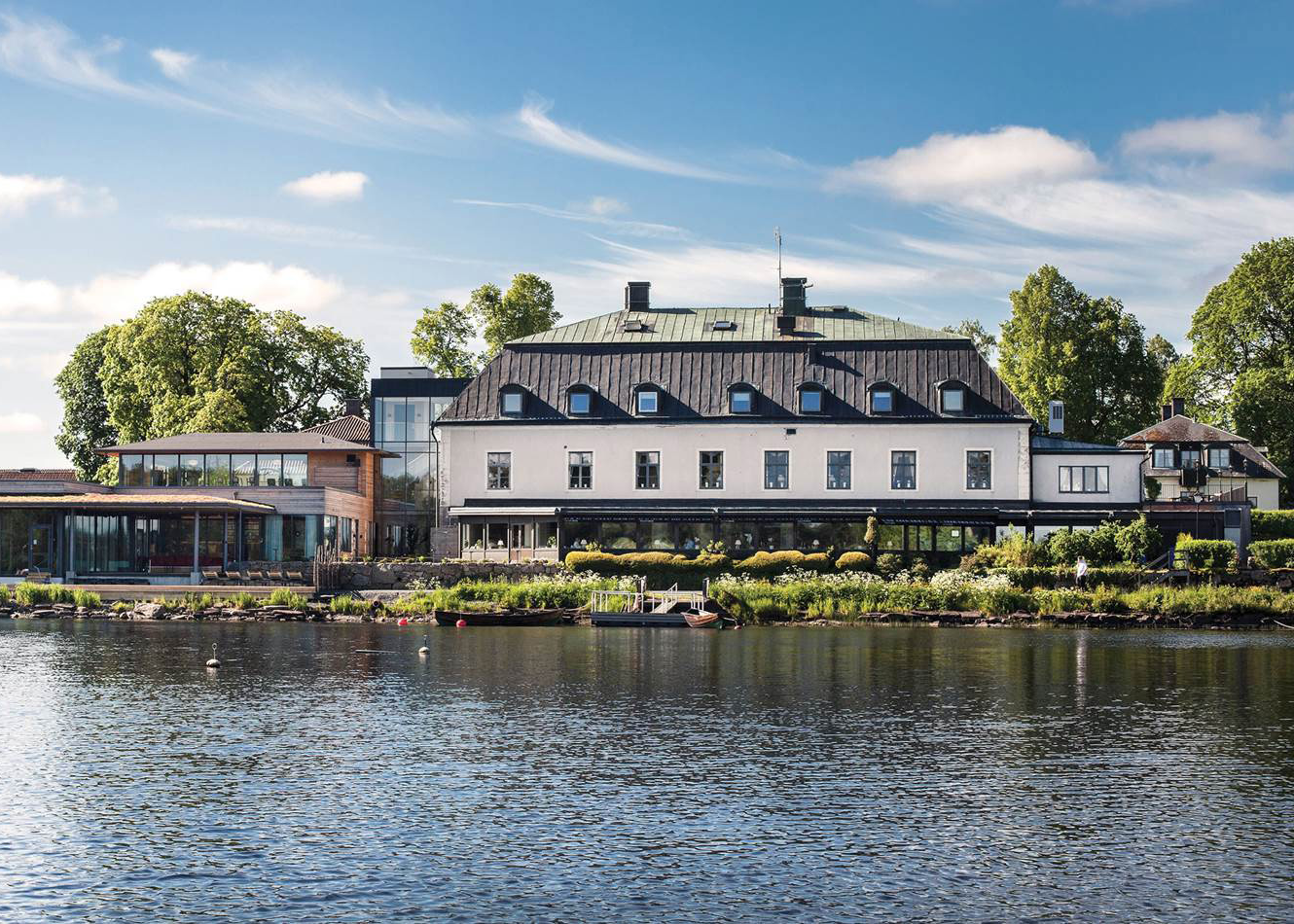 Stort hotell med svart tak och vita väggar ligger vid en sjö, omgiven av grönska och träd under blå himmel.