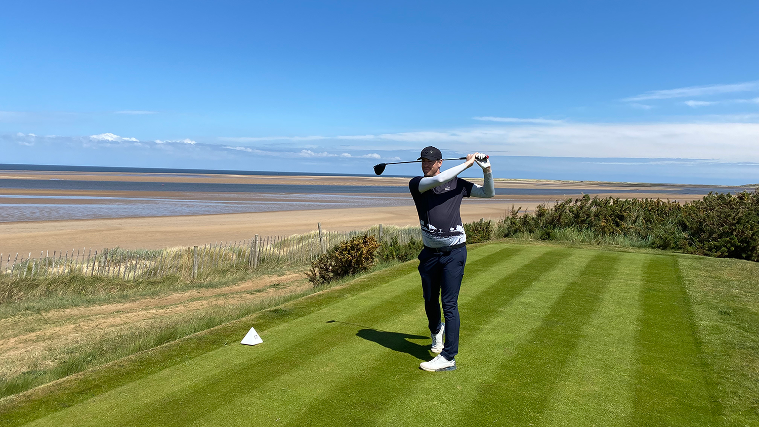 En golfspelare svingar en klubba på en välklippt tee med havet och en sandstrand i bakgrunden under klarblå himmel.