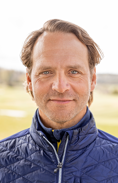 Man med mellanlångt brunt hår bär blå quiltad jacka och står utomhus med suddig bakgrund av gräs och träd.