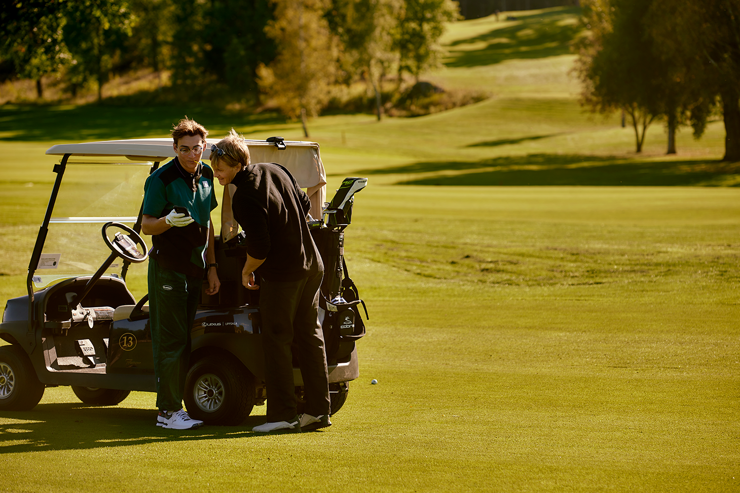 Två personer står vid en golfbil på en solig golfbana, en håller i en mobiltelefon och de tittar på skärmen.